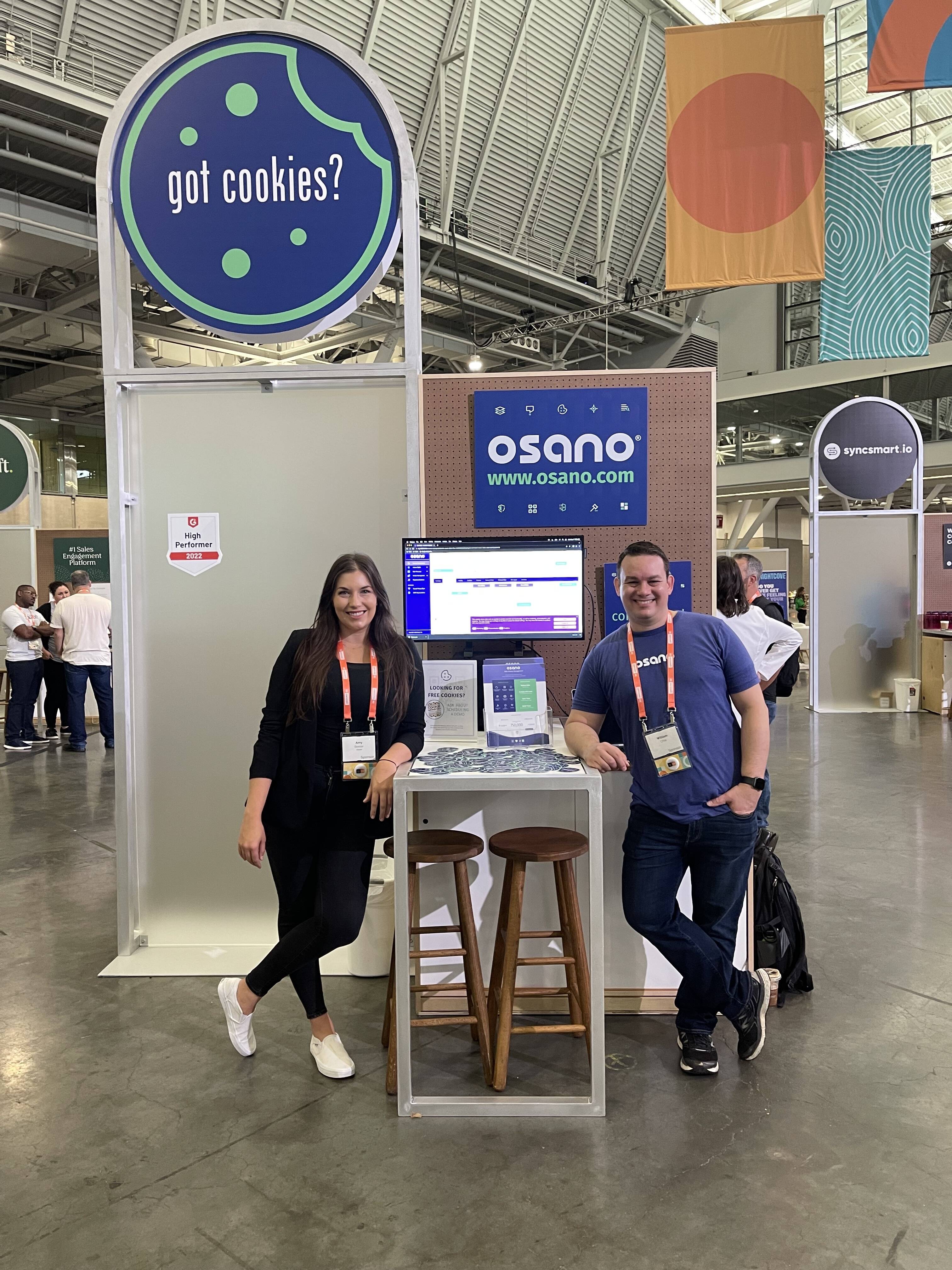 Two of Osano's team members lean on the company's booth at INBOUND. Behind them, a sign that says, "Got Cookies?" The booth -- one high-top table and two stools, has a bunch of stickers on it, a monitor with Osano's product, and some sales assets for attendees.